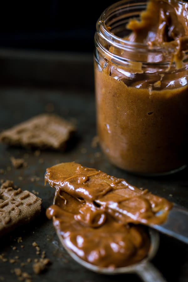 Speculoos spread on a knife and spoon, with a glass jar and biscoff biscuits in the background.
