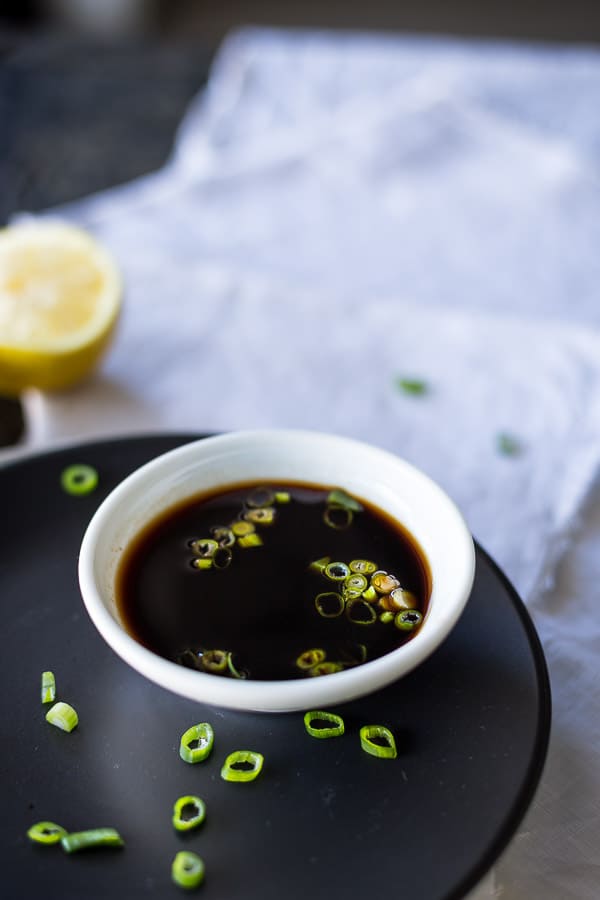 Ponzu sauce in a dipping bowl on a black plate with lemon in the background.