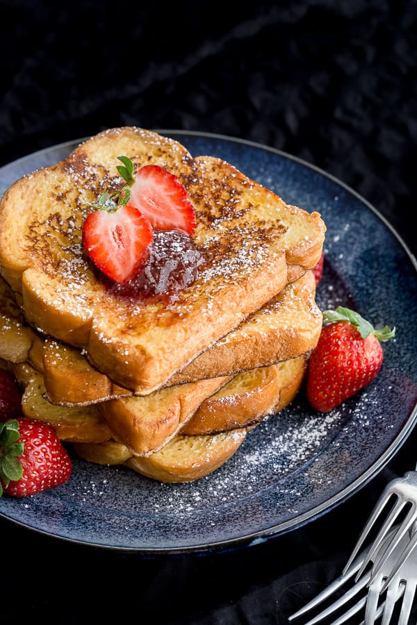A stack of freshly cooked french toast slathered with strawberry jam and topped with fresh strawberries.