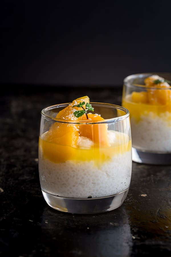 Two glasses of sago pudding showing the creamy white sago layer topped with yellow mango slices in syrup. 