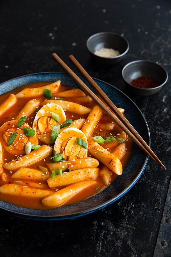 Bowl of Tteokbokki with boiled eggs, chopsticks and pinch bowls of sesame seeds and Korean chilli flakes.