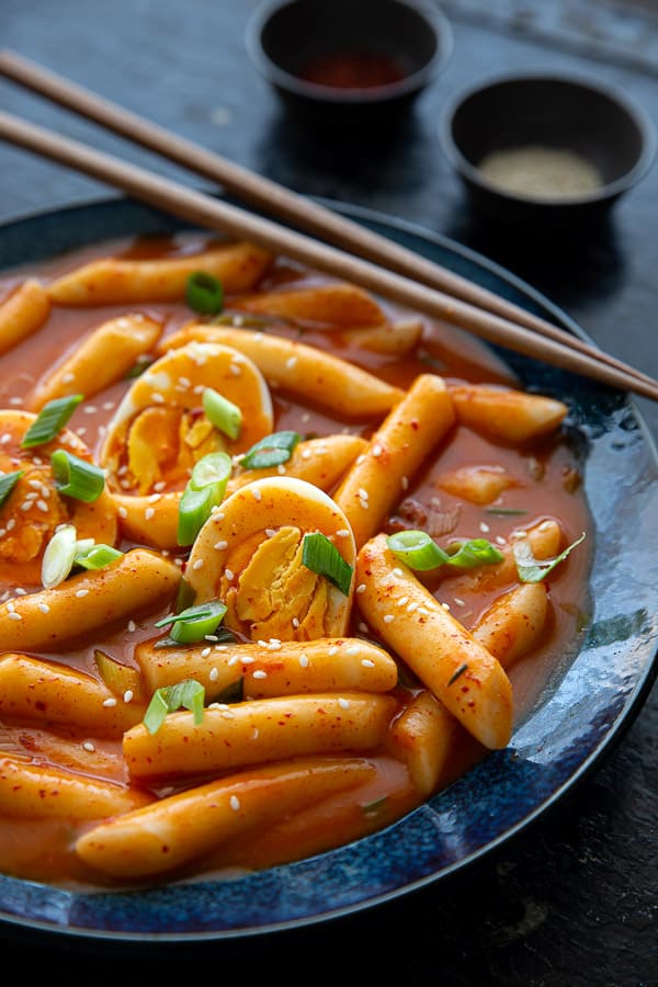 Bowl of tteokbokki with boiled eggs, chopsticks and pinch bowls of sesame seeds and Korean chilli flakes.