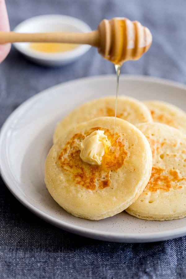 Honey being poured over fresh crumpets with butter on plate.