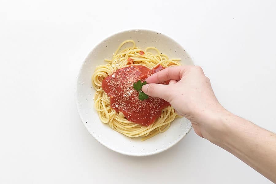 Bowl of pasta topped with napoli sauce and hand placing a parsley leaf.