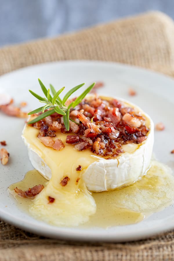 A melted camembert cheese on a plate topped with bacon and rosemary.