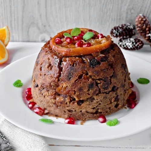 Christmas pudding on a white plate topped with candied fruit and pomegranate seeds.