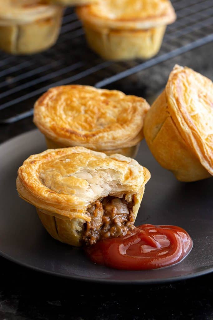 Mini party pies with beef filling, served on black plate with ketchup.