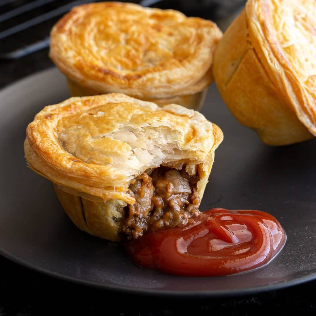 Beef pie on a plate with tomato sauce and more pies in the background.