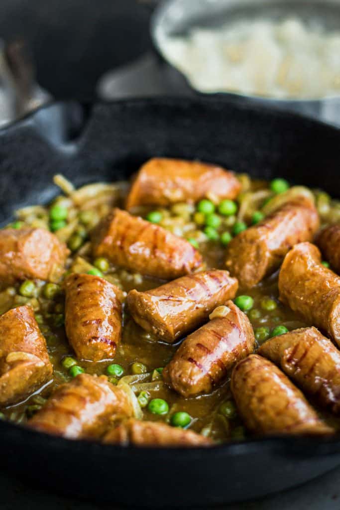 Chopped up sausages cooking in a pan with peas, onions and curry gravy.