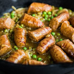 Close up shot of sausages cooking in a curry gravy with peas and onions.