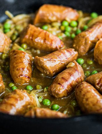 Close up shot of sausages cooking in a curry gravy with peas and onions.