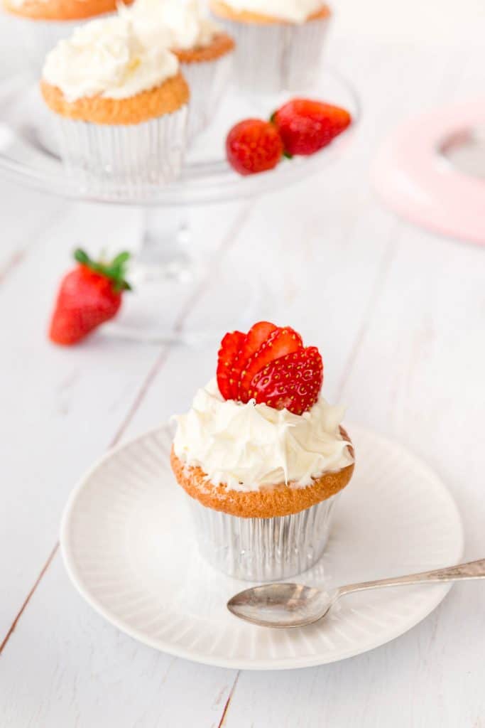 Cupcake on a plate, next to more cupcakes on a cake stand and a few fresh strawberries. 
