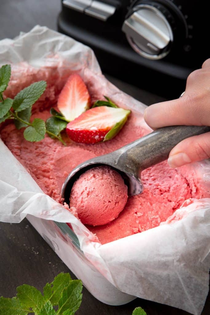 Using an ice cream scoop to scoop strawberry ice cream out of the dish.