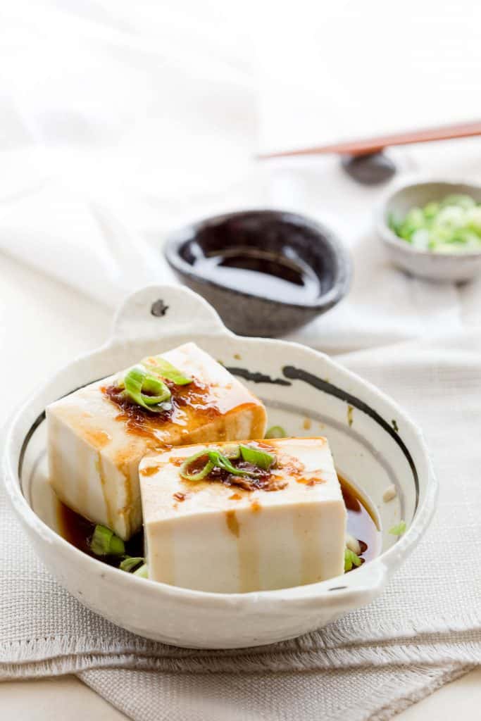 Two slices of yudofu in a bowl drizzled with sauce and garnished with spring onion.