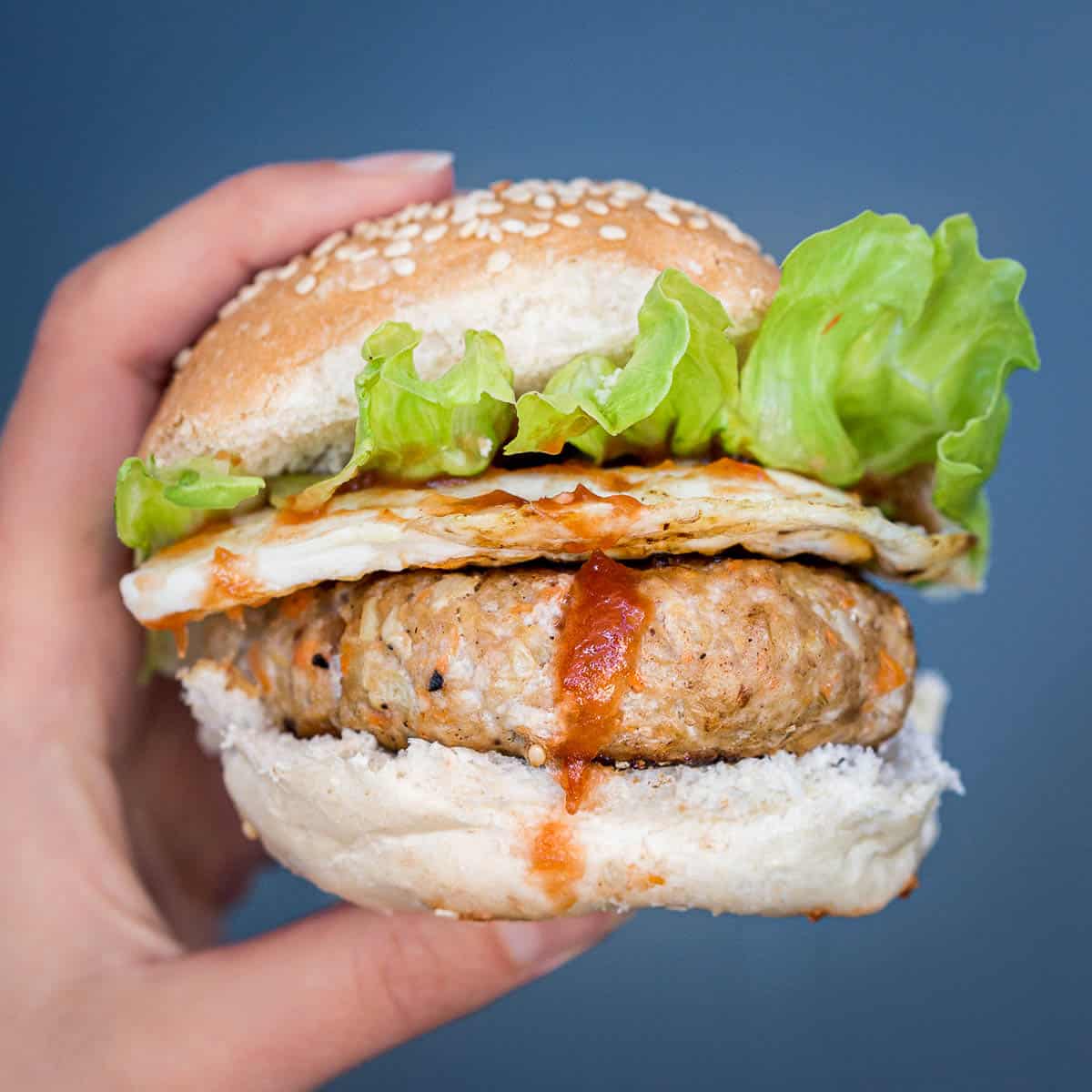 Hand holding a small pork burger with egg and lettuce on a sesame seed bun.