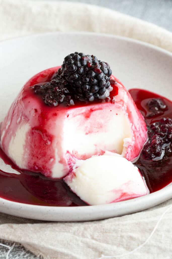 Blancmange on a plate drizzled with blackberry coulis, a portion has been sliced out with a spoon.