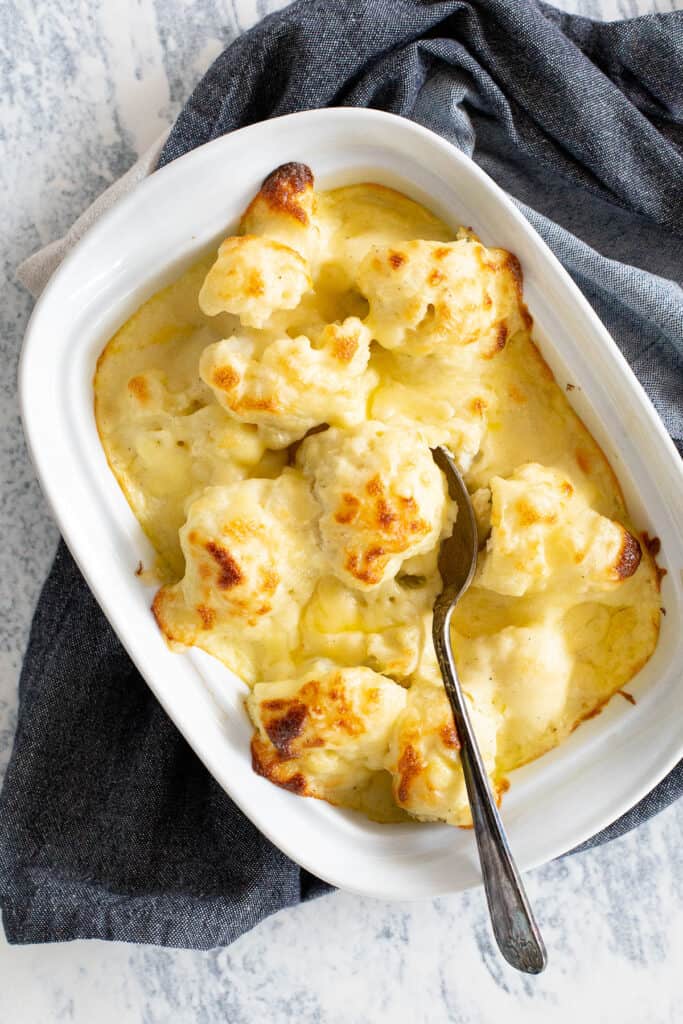 A metal spoon rests in a big baking dish filled with fresh cauliflower cheese.