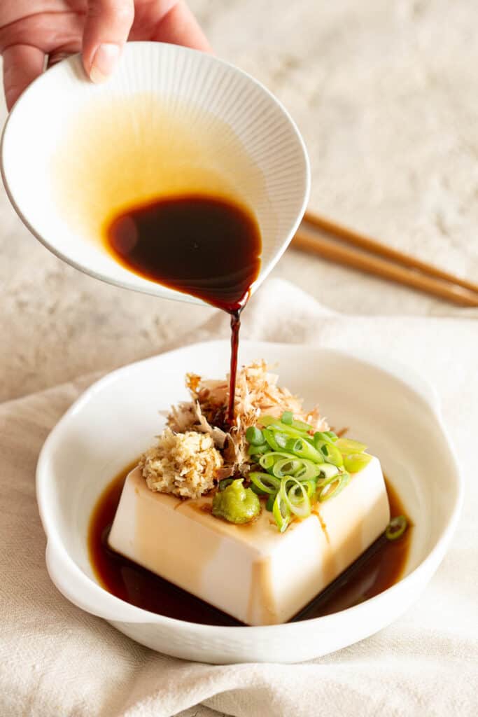 A hand is pouring a small dish of soy sauce over the Japanese cold tofu.