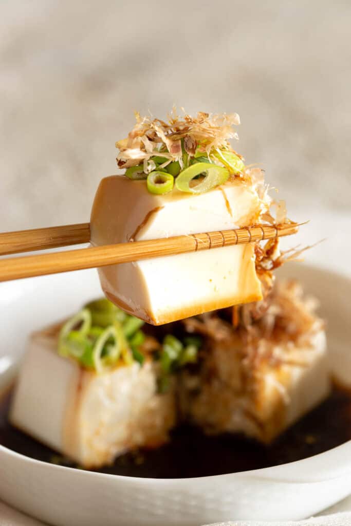 A pair of wooden chopsticks hold up a piece of silken tofu topped with bonito and spring onion.