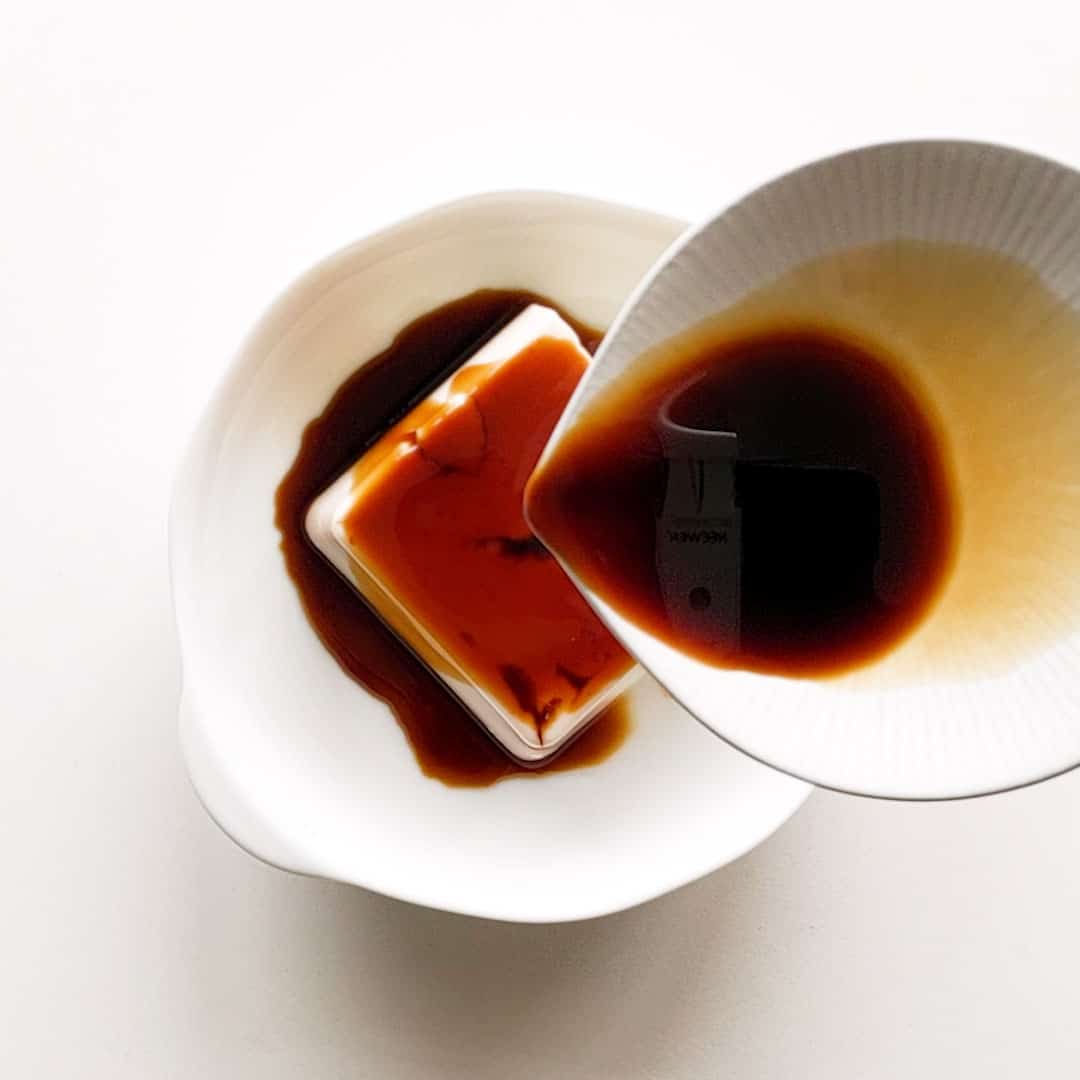A white dish pouring soy sauce over a square piece of silken tofu.