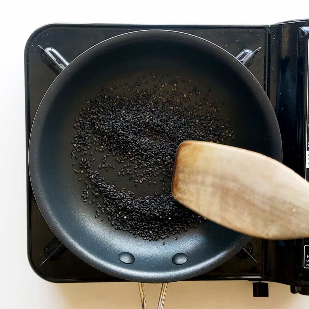 A wooden spatula is mixing black sesame seeds in a frying pan.