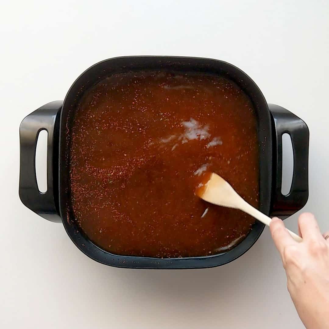 Stirring red broth in a shallow frying pan with a wooden spoon.