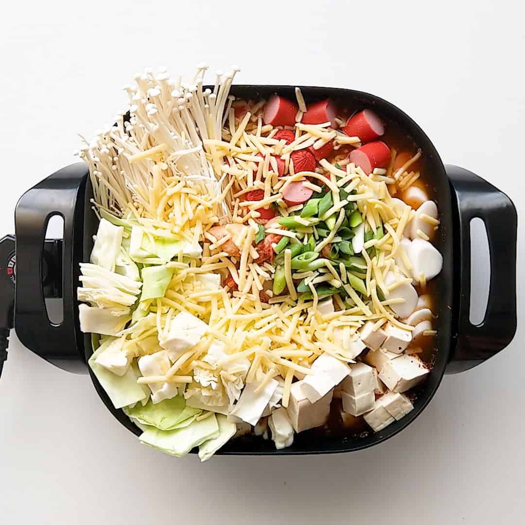 Korean army stew ingredients in an electric frying pan with shredded cheese scattered over the top.
