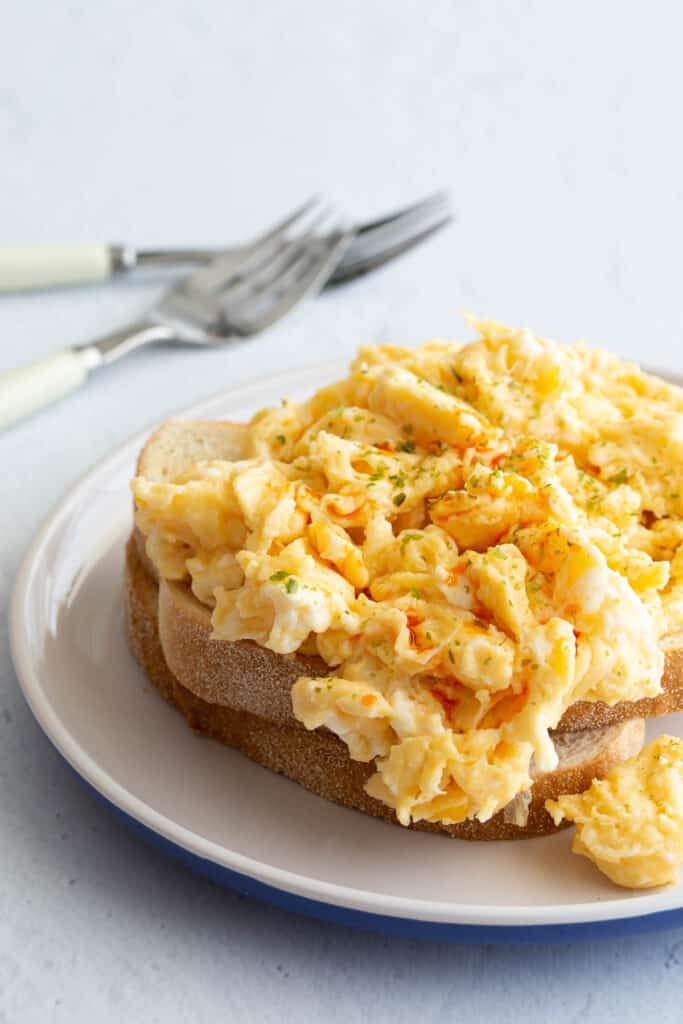 Miso butter scrambled eggs are topped with aonori and chilli sauce on a white place with forks in the background.