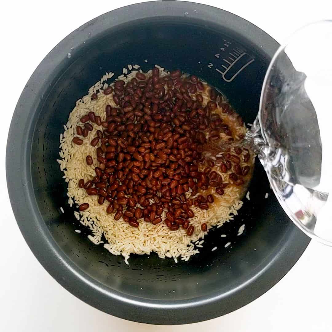 Pouring water over sticky rice and cooked red beans in a rice cooker bowl.