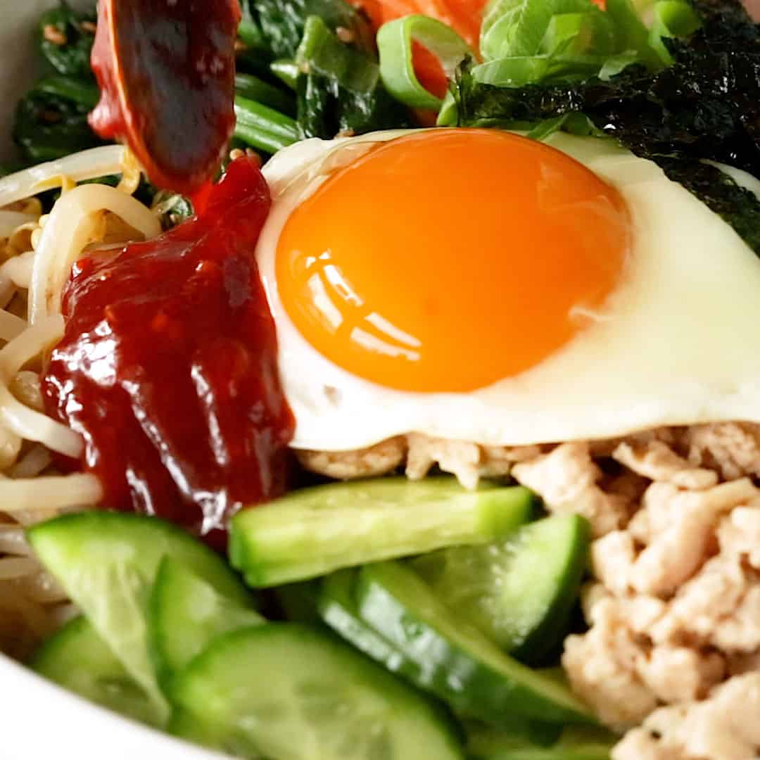 Pouring bibimbap sauce over a bowl of cooked chicken, vegetables, and sunny fried egg.