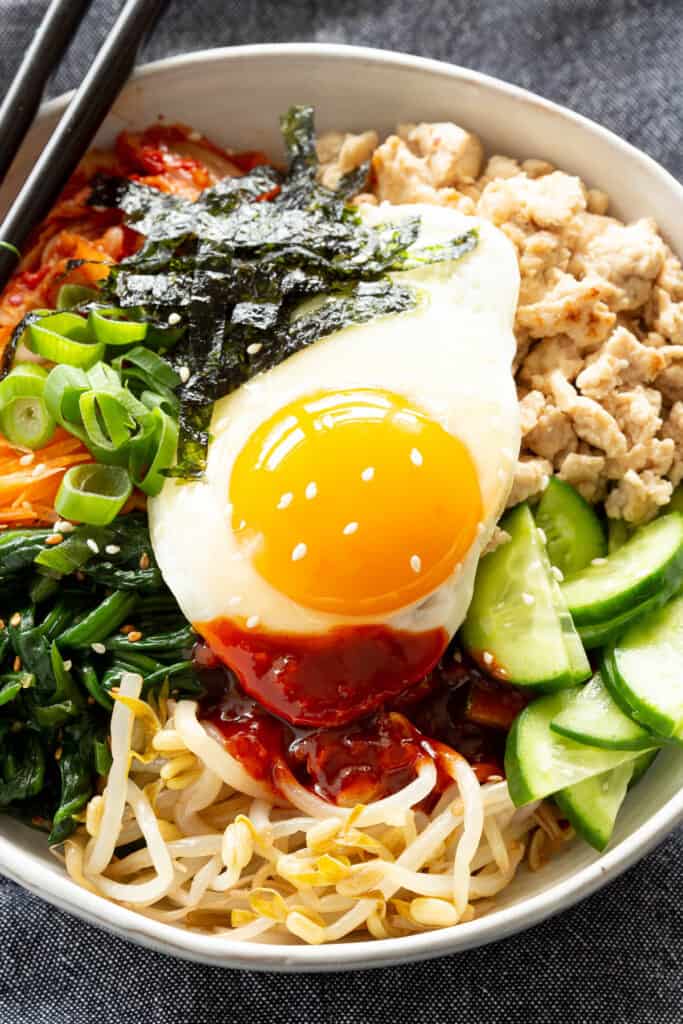A bright and colourful bowl of Chicken Bibimbap sits on a dark blue background with chopsticks.