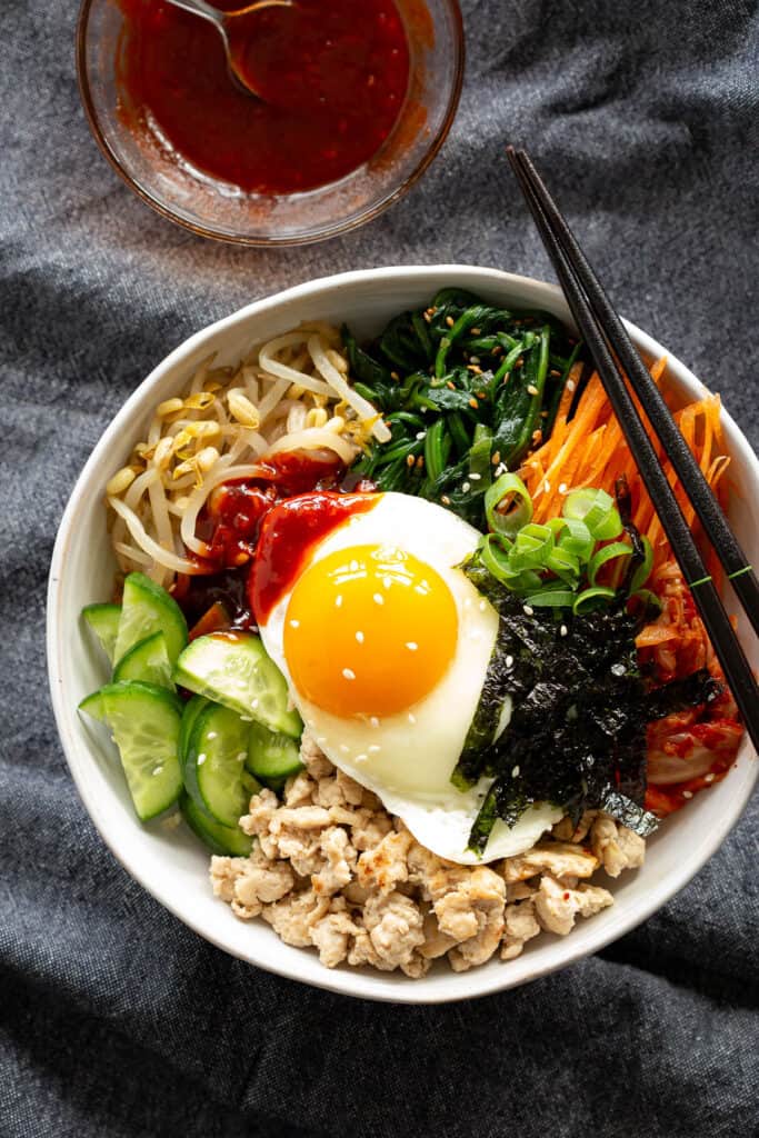 A Korean rice bowl in a white dish topped with chicken, egg, bean sprouts, spinach, carrot and seaweed.