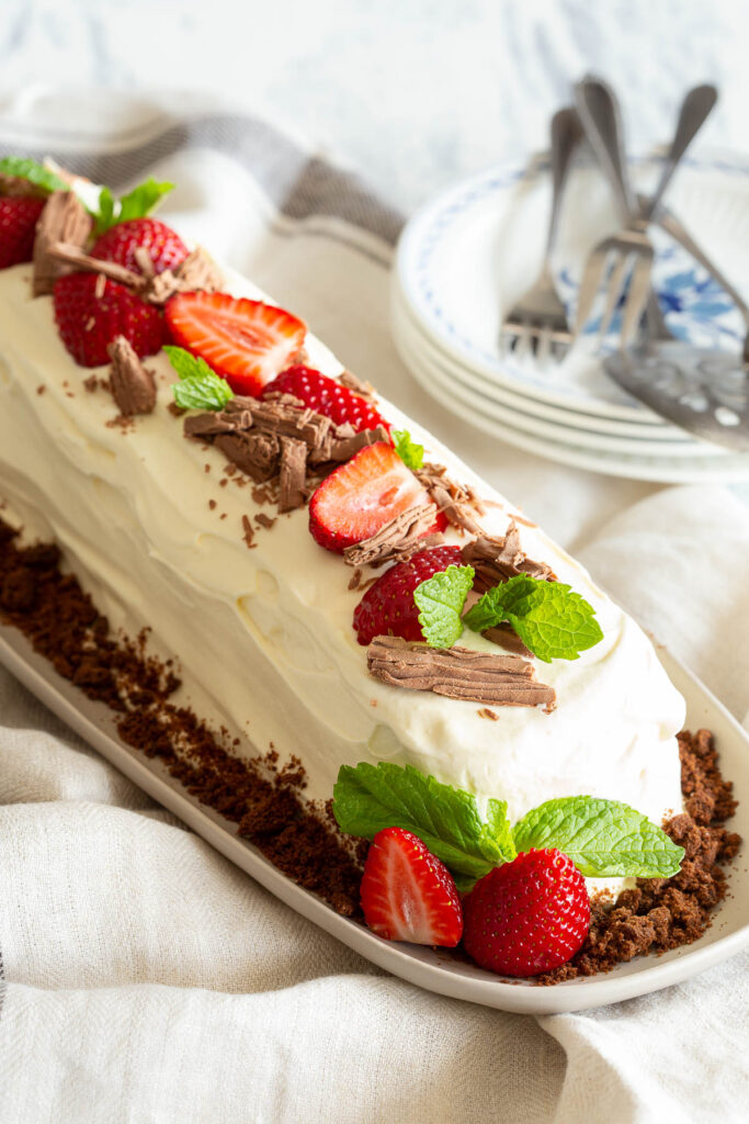 A chocolate ripple cake sits proud on a plate, with a garnish of biscuit crumbs, strawberry and mint.