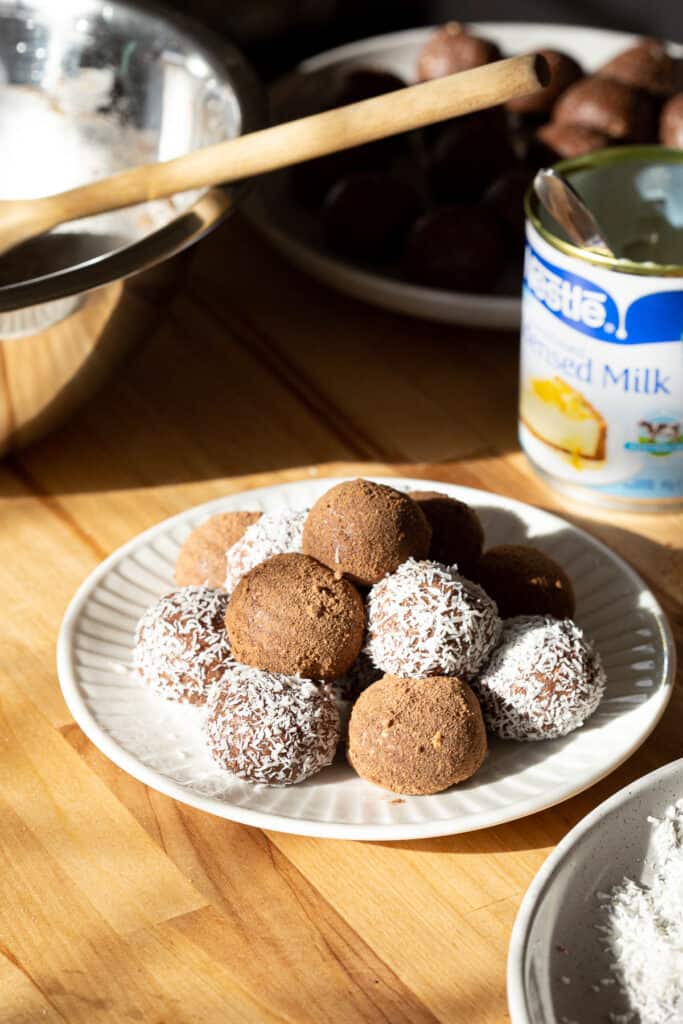 A stack of chocolate coconut balls sits beside a tin, bowl and another plate.
