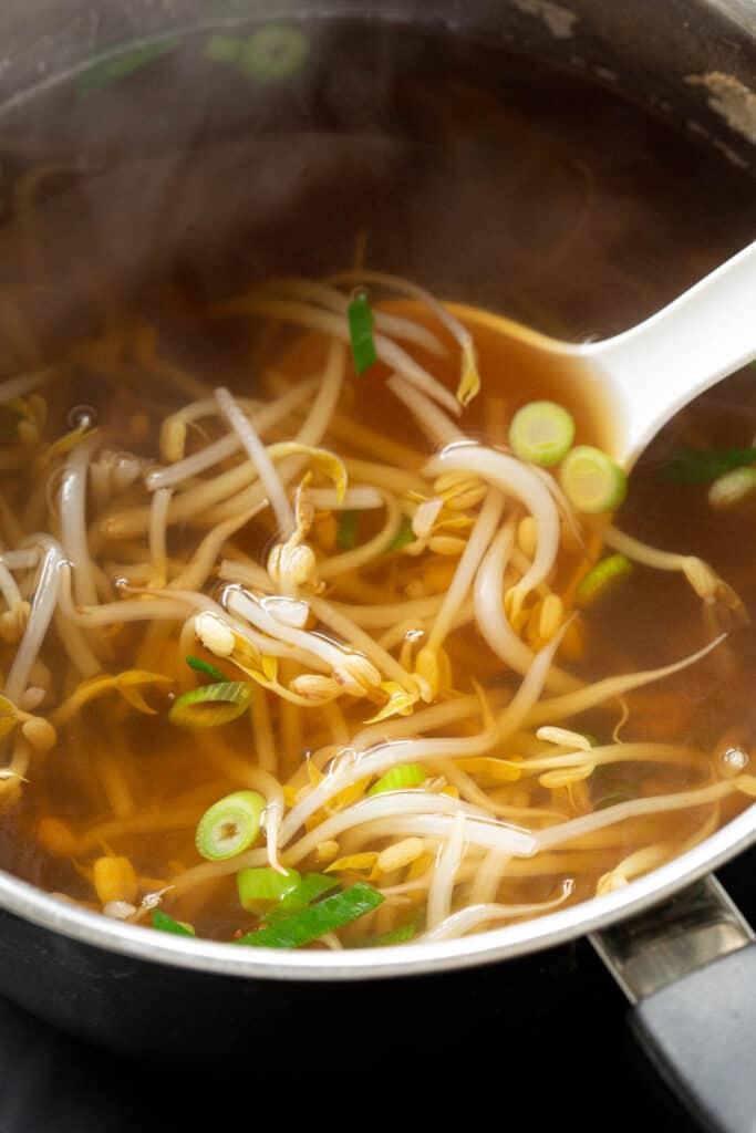 A ladle stirs the bean sprouts around the soup in a large pot.