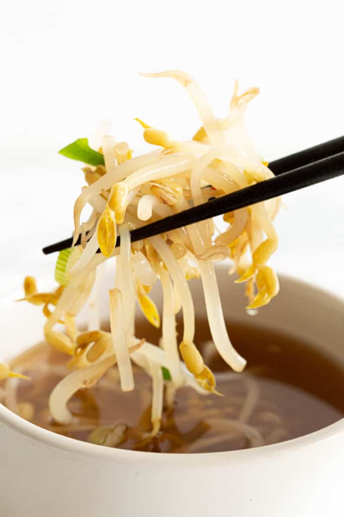Chopsticks holding up bean sprouts above a bowl of soup.