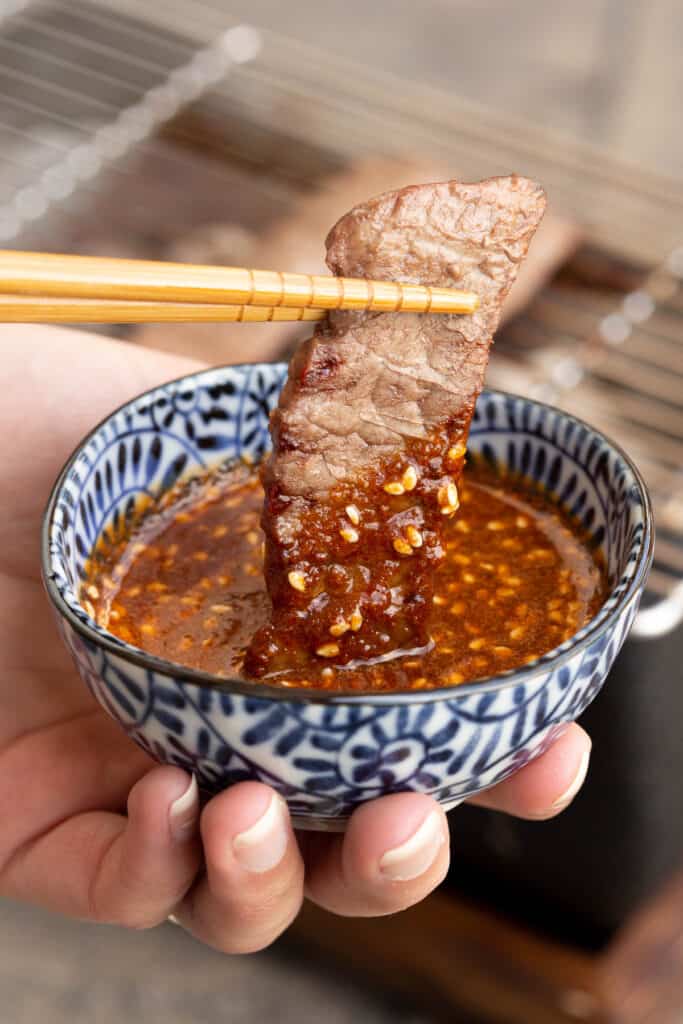 A piece of grilled meat is dipped into a small bowl of yakiniku sauce.