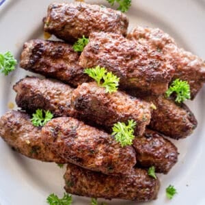 A stack of grilled cevapi on a white plate with parsley garnish.