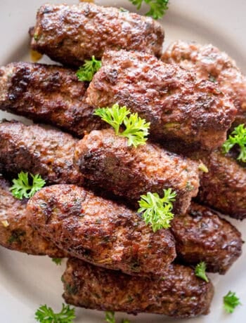 A stack of grilled cevapi on a white plate with parsley garnish.