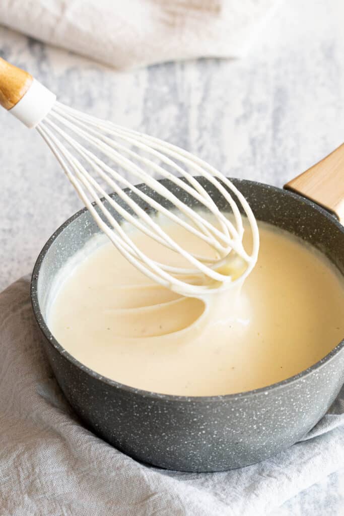 A whisk scoops up silky smooth white sauce out of the pan.