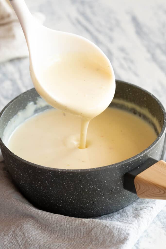 A ladle pours creamy white sauce into a grey saucepan.