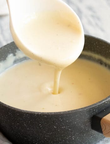 A white spoon pours smooth white sauce into a saucepan with wooden handle.