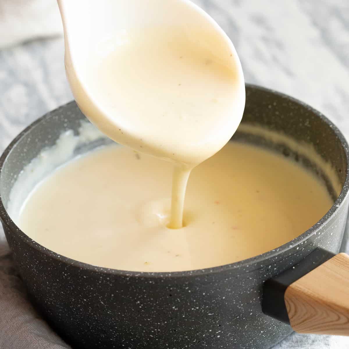 A white spoon pours smooth white sauce into a saucepan with wooden handle.