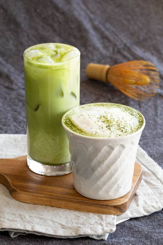 A hot and iced matcha latte sit on a small wooden serving board with a chasen in the background.