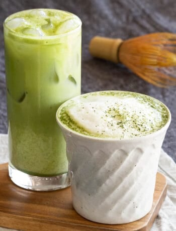 Two matcha lattes sit on a small wooden serving board with a chasen in the background.