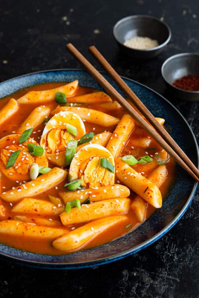 A dark bowl filled with tteokbokki and boiled eggs in a rich red sauce. Garnished with sesame seeds and green onions.