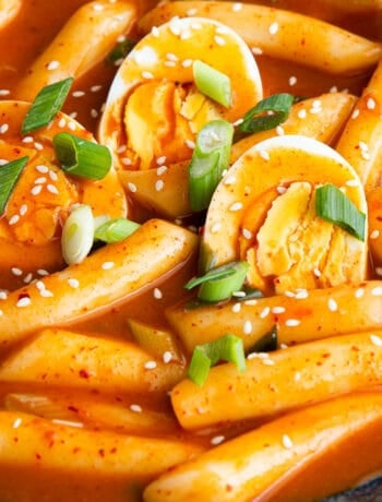 Close up shot of tteokbokki rice cakes and boiled eggs in a bright red and glossy sauce with toasted sesame seeds and spring onion as garnish.