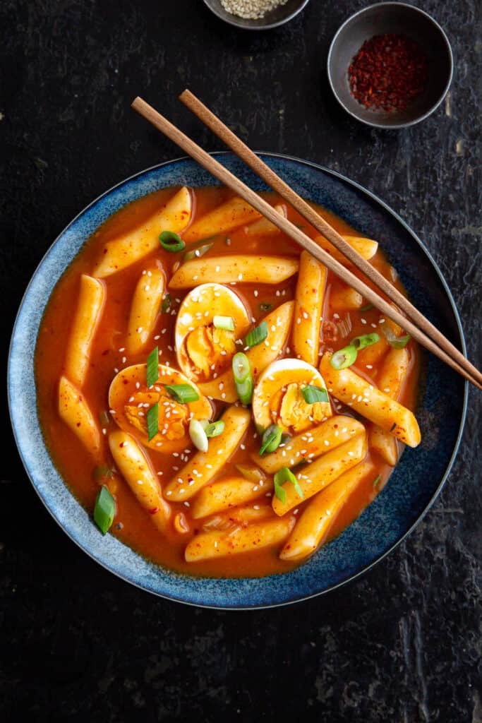 Bright red tteokbokki in a blue bowl. A pair of chopsticks rests across the edge.
