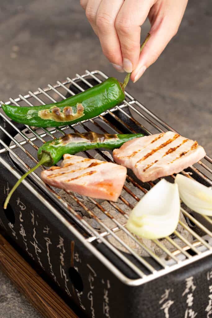 A hand turns over a grilled green pepper on the hibachi.