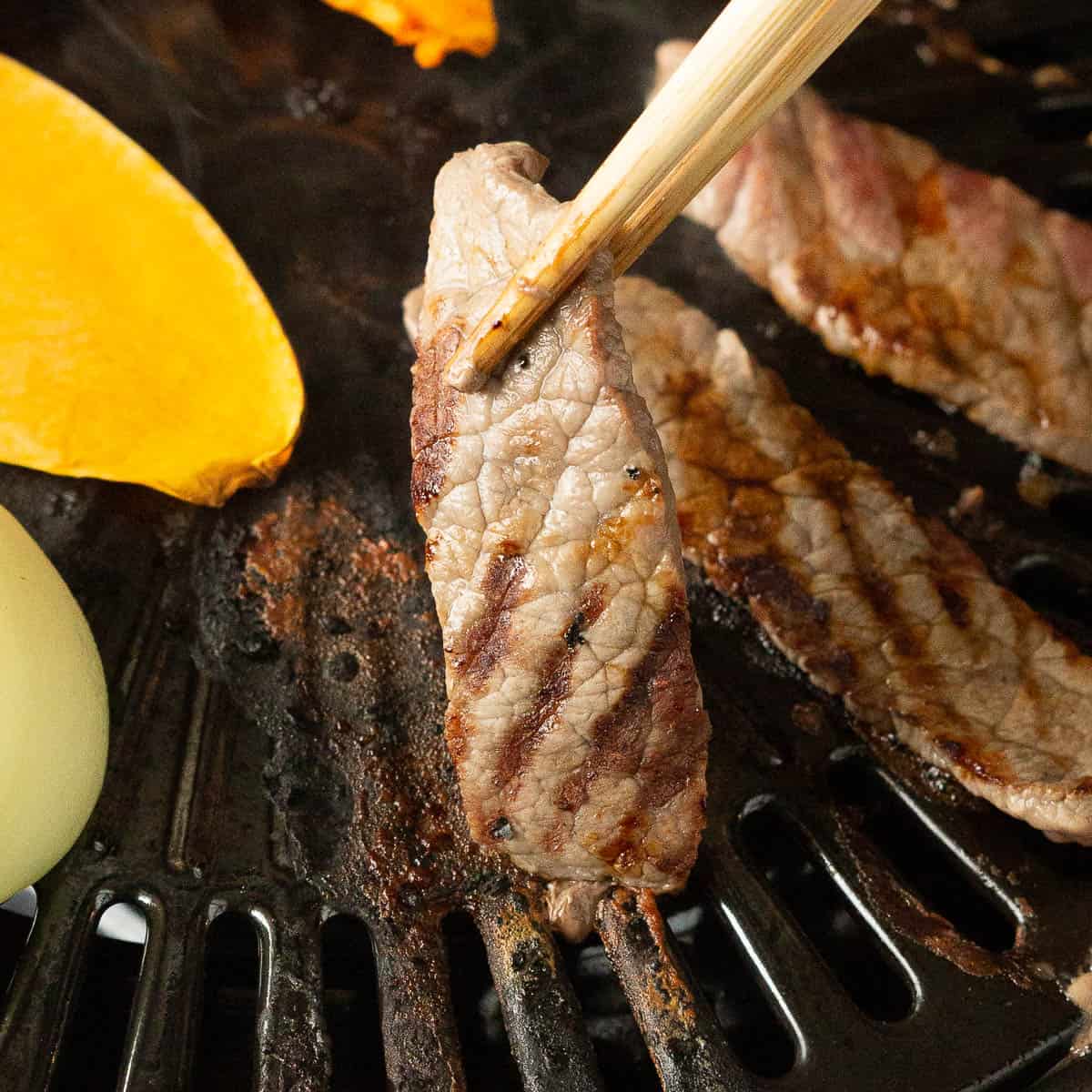 A thin slice of beef with grill marks being picked up by chopsticks.